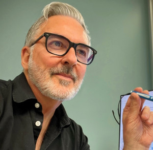 Murray Bays wearing black glasses and shirt, holds a pen thoughtfully.